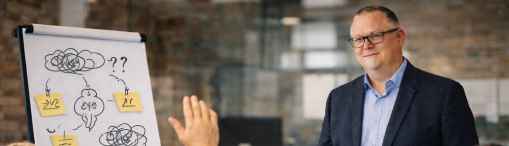 Moderationssituation in einem Arbeitsumfeld. Alexander Vollberg steht aufmerksam vor einer Gruppe von Teilnehmenden. Die Atmosphäre wirkt konzentriert und sachlich. Es geht um Austausch, Klärung und gemeinsames Verstehen organisatorischer Herausforderungen.